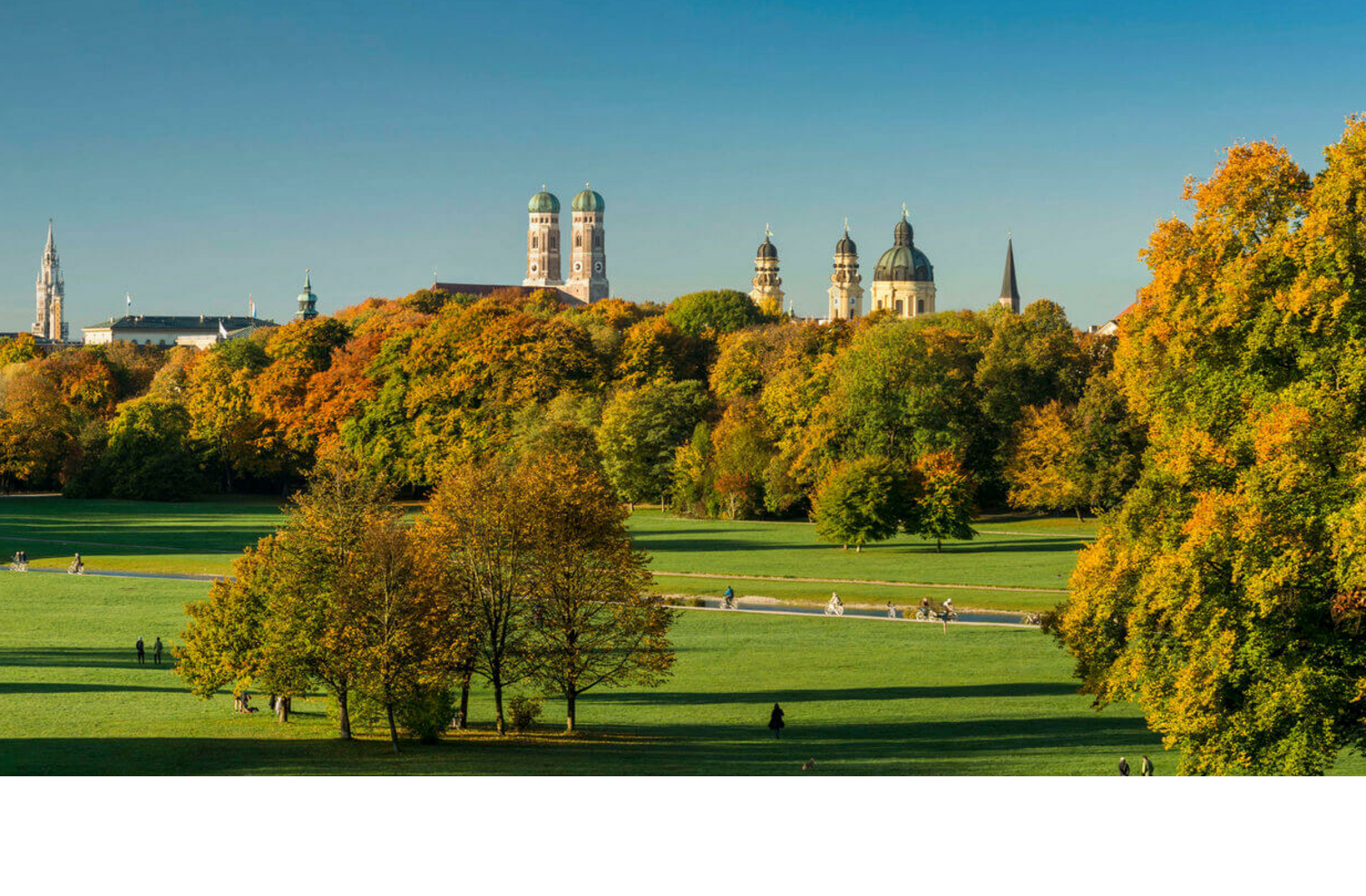Spacious park with green meadows, colorful autumn trees and a city silhouette in the background under a clear sky.