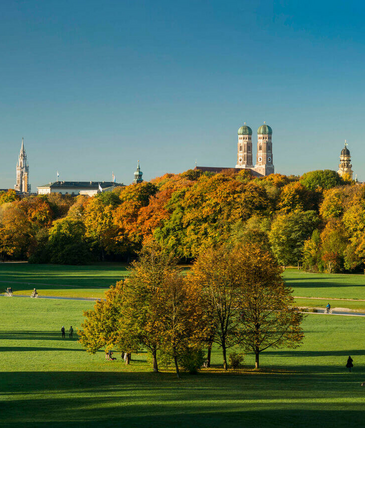 Panorama im Englischen Garten Weitläufiger Park mit grünen Wiesen, bunten Herbstbäumen und einer Stadtsilhouette im Hintergrund unter klarem Himmel.