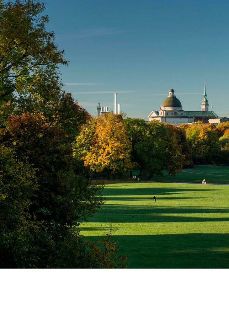 Panorama im Englischen Garten Weitläufiger Park mit grünen Wiesen, bunten Herbstbäumen und einer Stadtsilhouette im Hintergrund unter klarem Himmel.