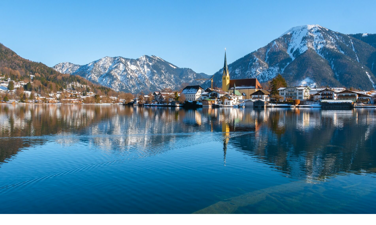 Tegernsee in München Idyllische Seelandschaft mit schneebedeckten Bergen, einem Dorf und einer Kirche am Ufer, reflektiert im ruhigen Wasser.