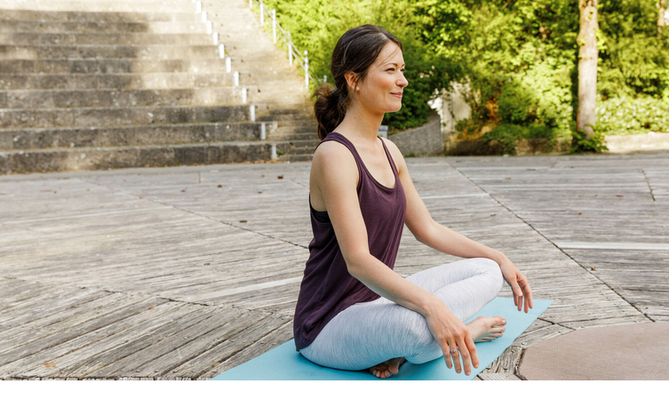 Woman meditating cross-legged on a yoga mat outdoors, surrounded by nature and stairs in the background.