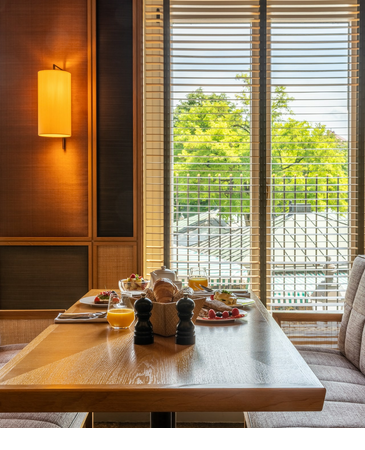 Breakfast table at the LOUIS Hotel with fresh food, juice and a view of green trees through large windows.