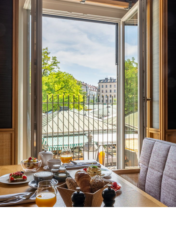 Fruehstueck LOUIS Hotel Muenchen Frühstückstisch im LOUIS Hotel mit Blick auf den Viktualienmarkt durch ein offenes Fenster.