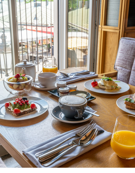 Breakfast table with coffee, juice, fruit and sandwiches at the LOUIS Hotel Munich, with a view of the market.