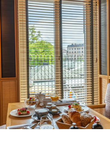 Cozy breakfast corner in the LOUIS Hotel with a laid table, view of the cityscape through shutters.