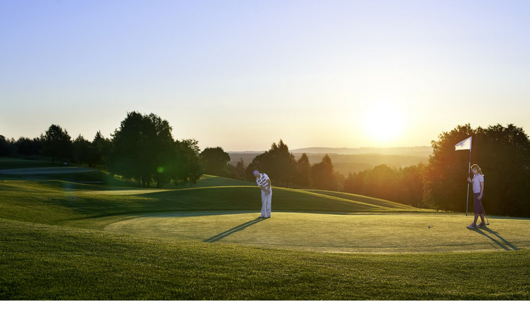 Golfers on a green course at sunset, one person holding the flag at the hole.
