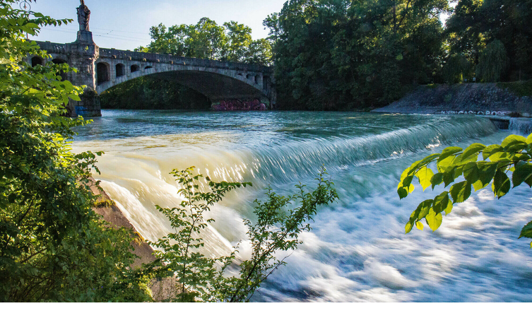 Isar on the Maximiliansbridge in Munich River with a small waterfall, a historic bridge above it, surrounded by lush greenery and trees.