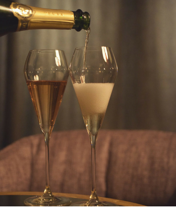 Two champagne glasses on a table, one being filled with champagne, a cozy armchair in the background.