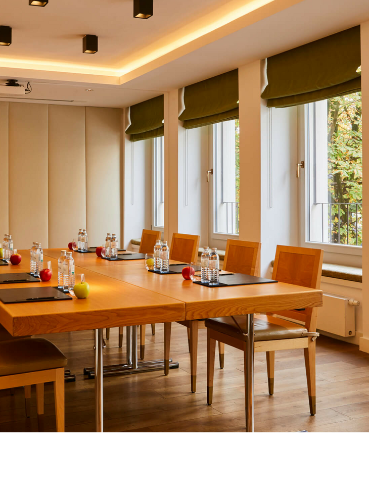 Meeting- und Tagungsraum Marketroom | LOUIS Hotel München Moderner Konferenzraum im LOUIS Hotel München mit Holzstühlen, Tisch, Wasserflaschen und Fenstern mit grünen Vorhängen.