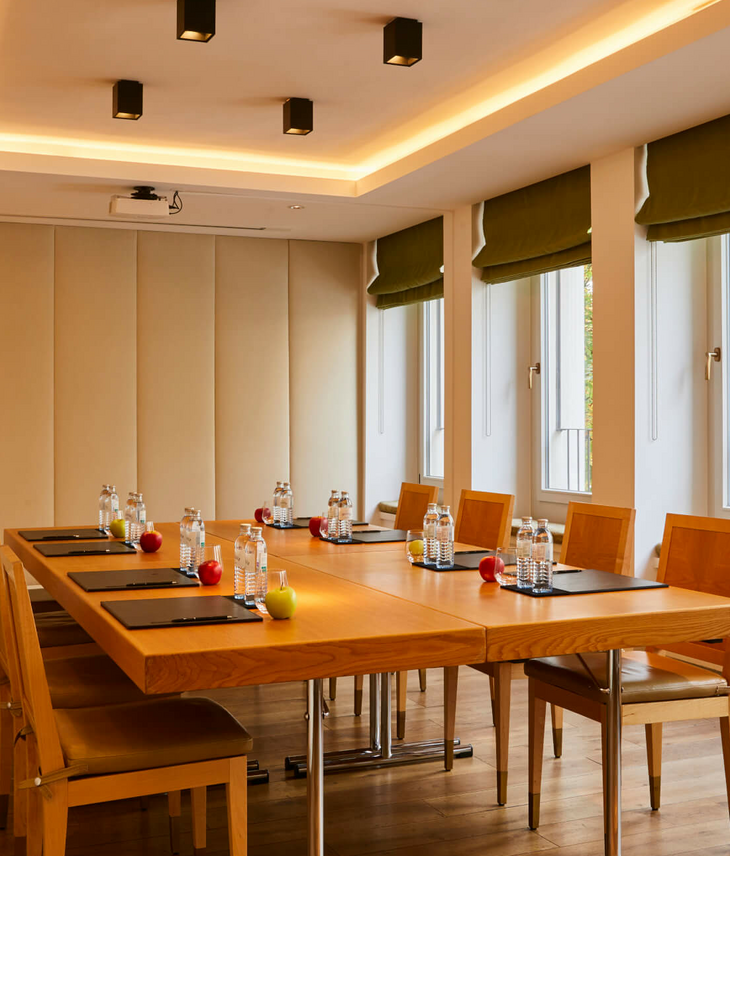 Modern conference room in the LOUIS Hotel Munich with wooden chairs, table, water bottles and windows with green curtains.