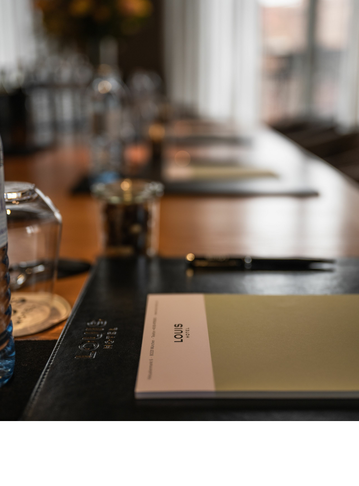 Nahaufnahme eines Notizblocks mit Louis Hotel München Logo, Wasserglas und Kugelschreiber