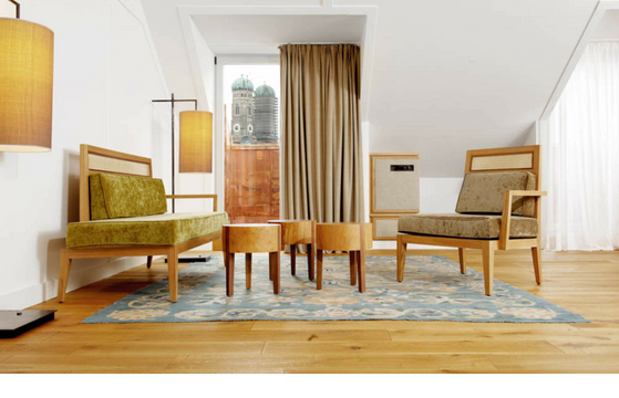 LOUIS Hotel München | Suite Helles Hotelzimmer mit stilvollen Holzmöbeln, Teppich und Blick auf Münchens Frauenkirche durch ein großes Fenster.