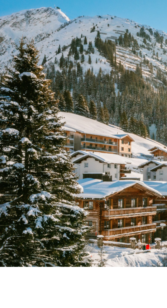 Verschneite Alpenlandschaft mit Sonne über Hotel Severin*s in Lech, umgeben von Bergen und Bäumen.