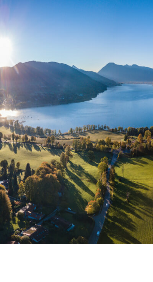 Aerial view of a lake with surrounding green meadows and mountains in the background at sunrise.