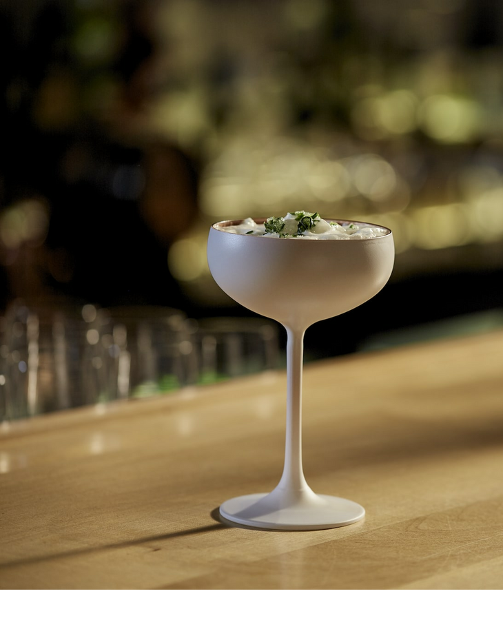 Ein eleganter Cocktail in einer weißen Schale auf einer Holztheke, unscharfer Hintergrund.