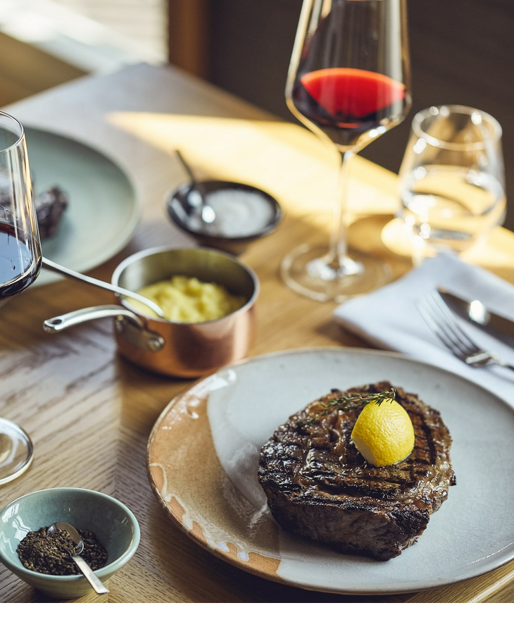 Steak with lemon, red wine glass, mashed potatoes and spices on a wooden table at the LOUIS Hotel Munich.