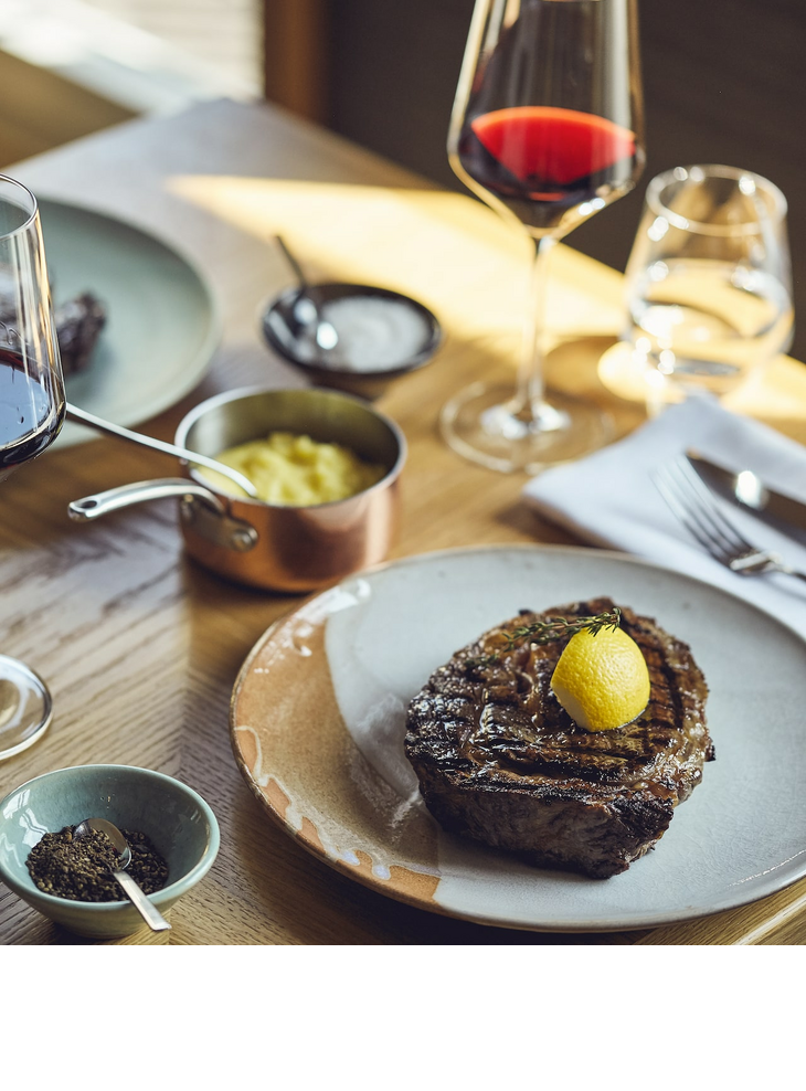 Steak with lemon, red wine glass, mashed potatoes and spices on a wooden table at the LOUIS Hotel Munich.