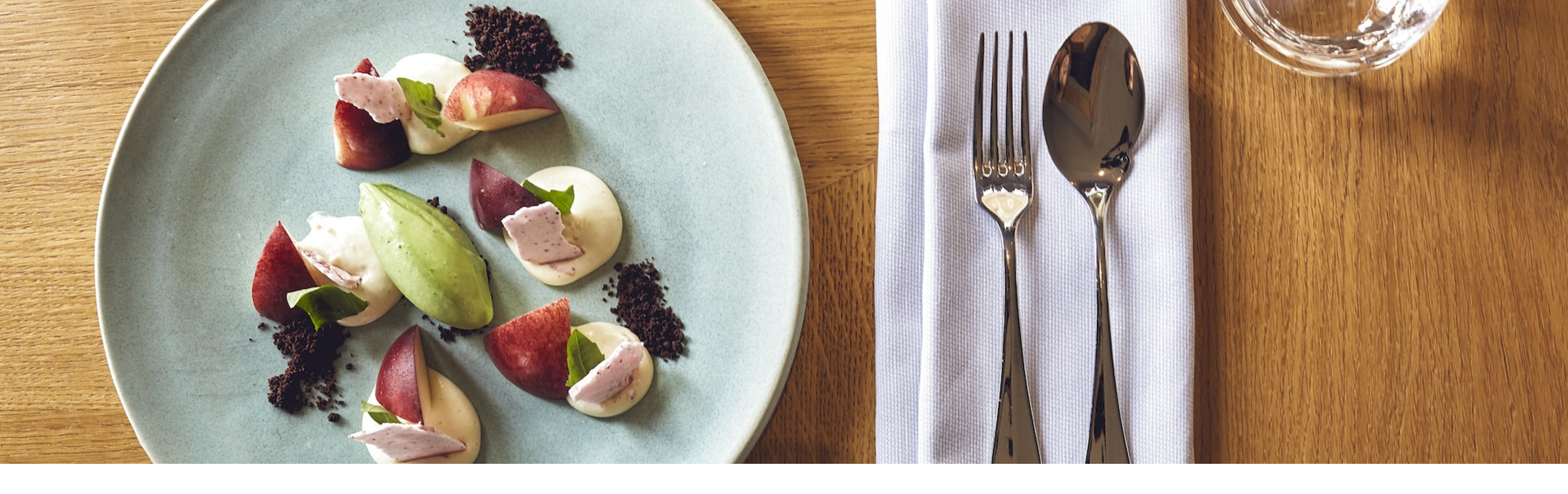 Elegant dessert on a blue plate with cutlery and glasses on a wooden table in the LOUIS Hotel Munich.