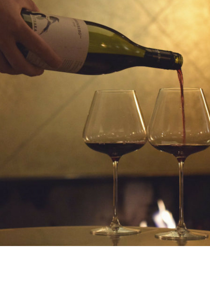 Hand pours red wine into two glasses in front of a cozy fireplace at the LOUIS Hotel Munich.