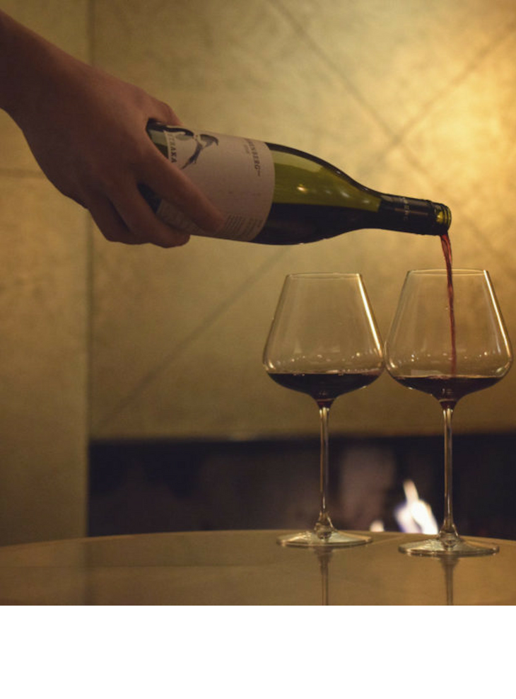 Hand schenkt Rotwein in zwei Gläser vor gemütlichem Kamin im LOUIS Hotel München ein.