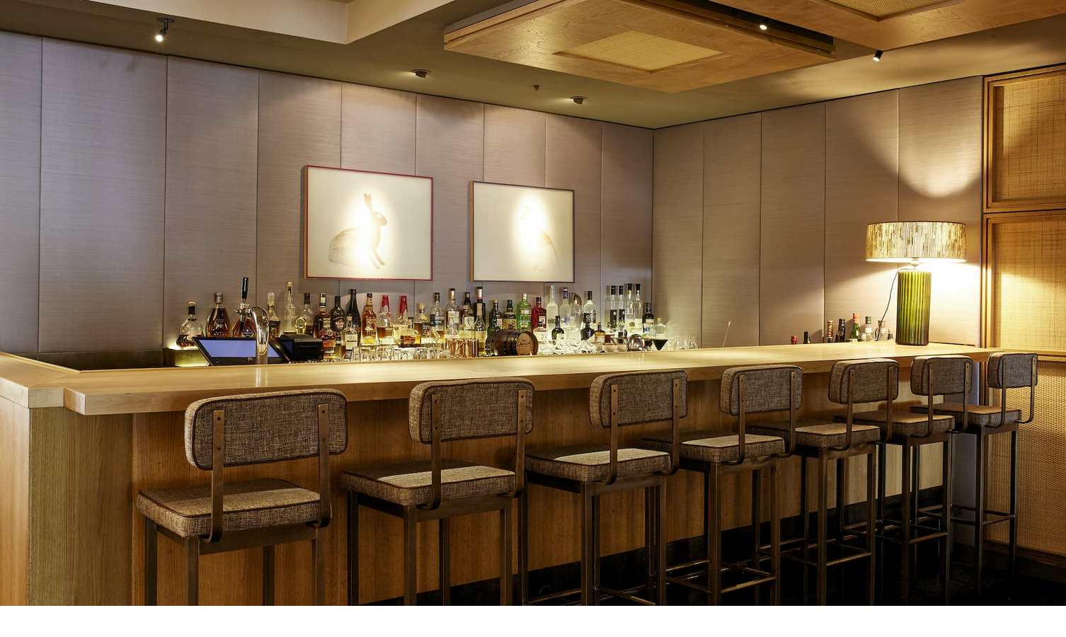 Elegant hotel bar with wooden counter, bar stools and stylish lighting at the LOUIS Hotel Munich.