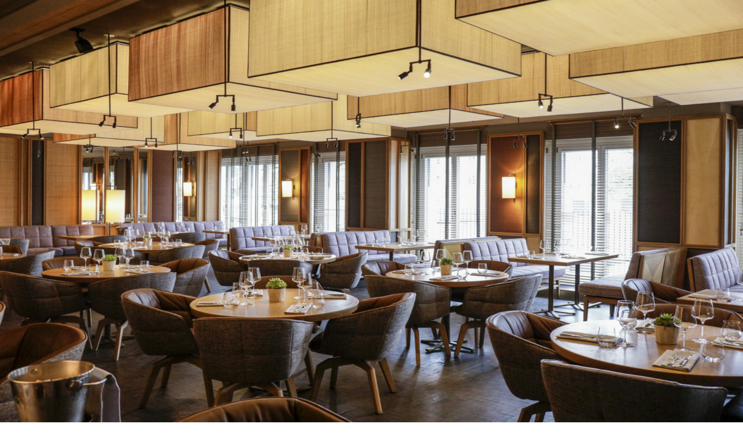 Elegant hotel restaurant with round wooden tables, upholstered chairs and modern lamps, daylight through windows.