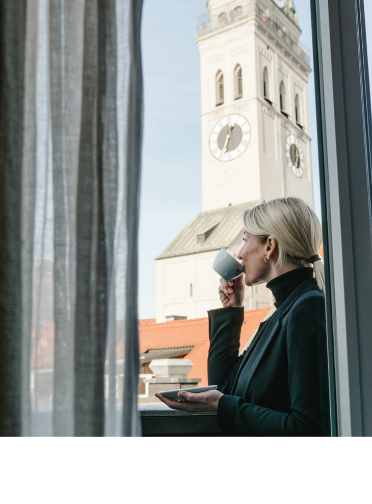 Frau trinkt einen Kaffee auf einem Balkon des LOUIS Hotels in München und blickt dabei auf die Stadt
