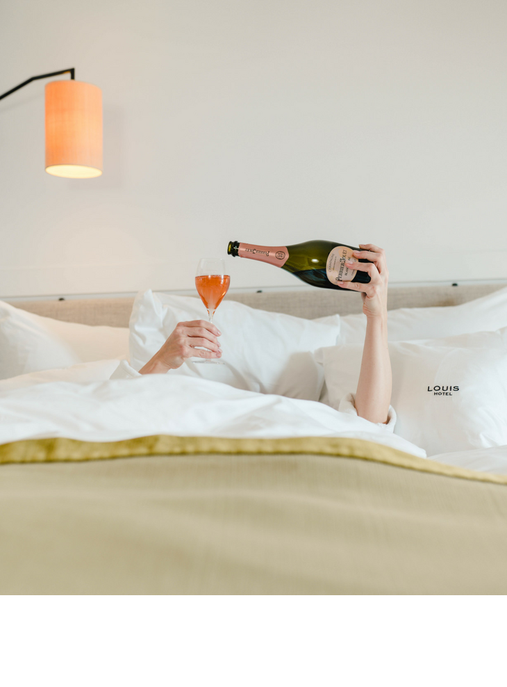 The LOUIS Hotel in München | Angebot Wochenende - Weekend rate Person im Bett gießt Sekt in ein Glas, umgeben von weißen Kissen im LOUIS Hotel München.