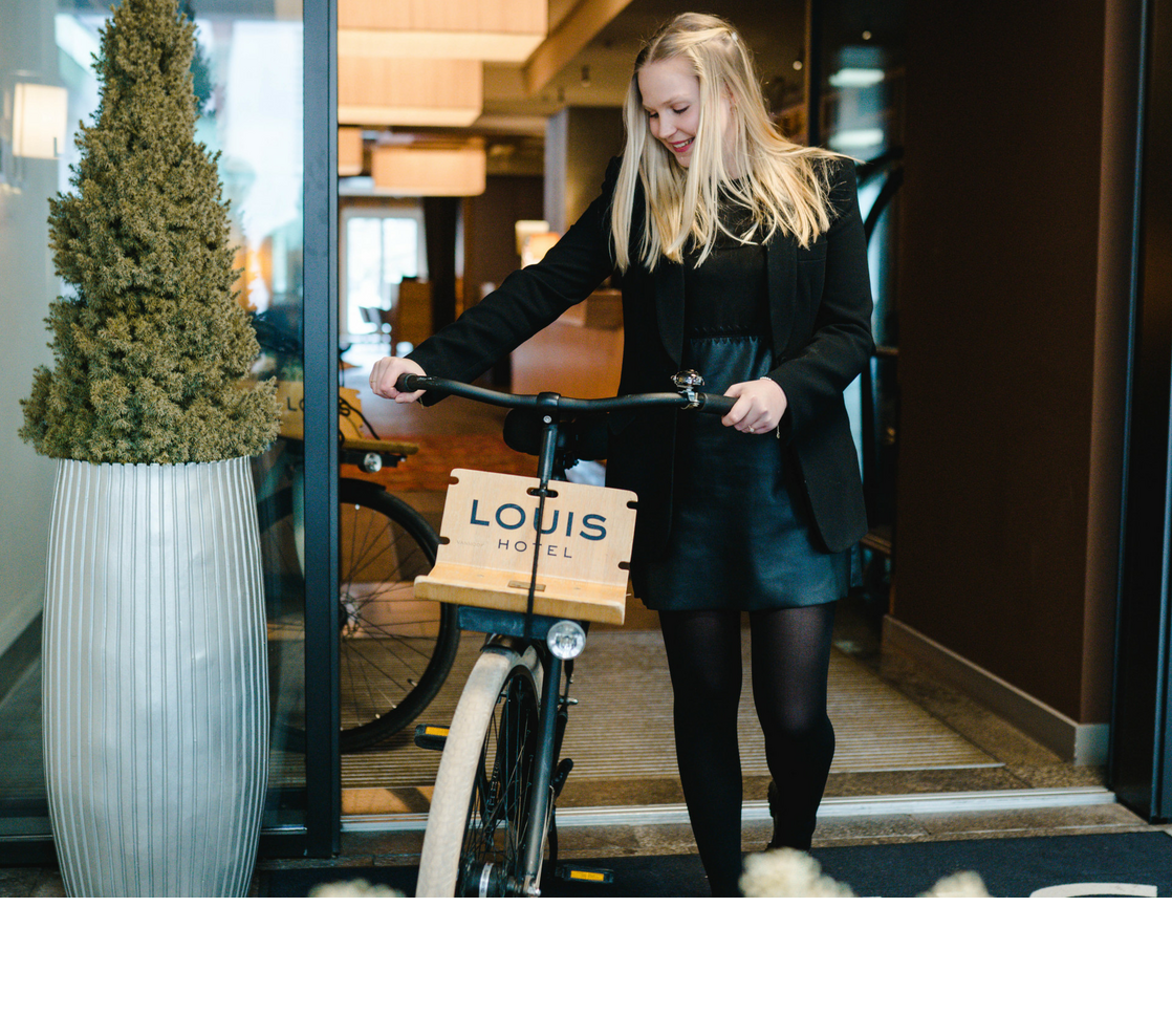 Rental bikes | The LOUIS Hotel Munich Woman with bicycle at the entrance of the LOUIS Hotel Munich, next to a large planter.