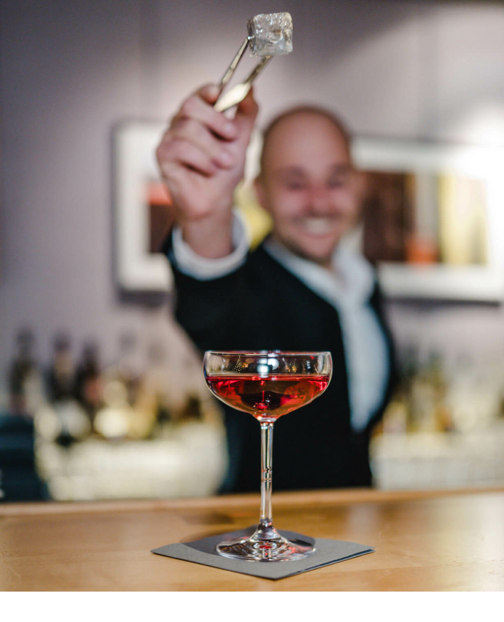 Bartender smiles and holds ice cube tongs over a red cocktail glass at the hotel bar.