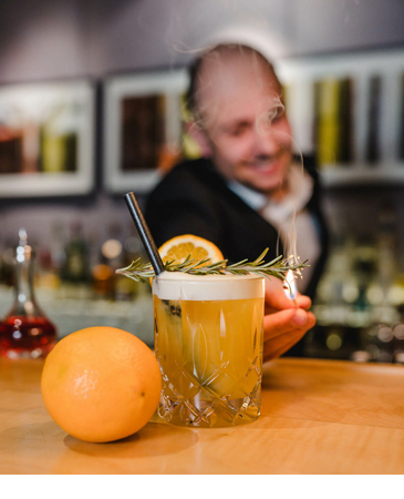 Ein Barkeeper serviert einen Cocktail mit Rosmarin und Orangenscheibe, daneben liegt eine Orange.